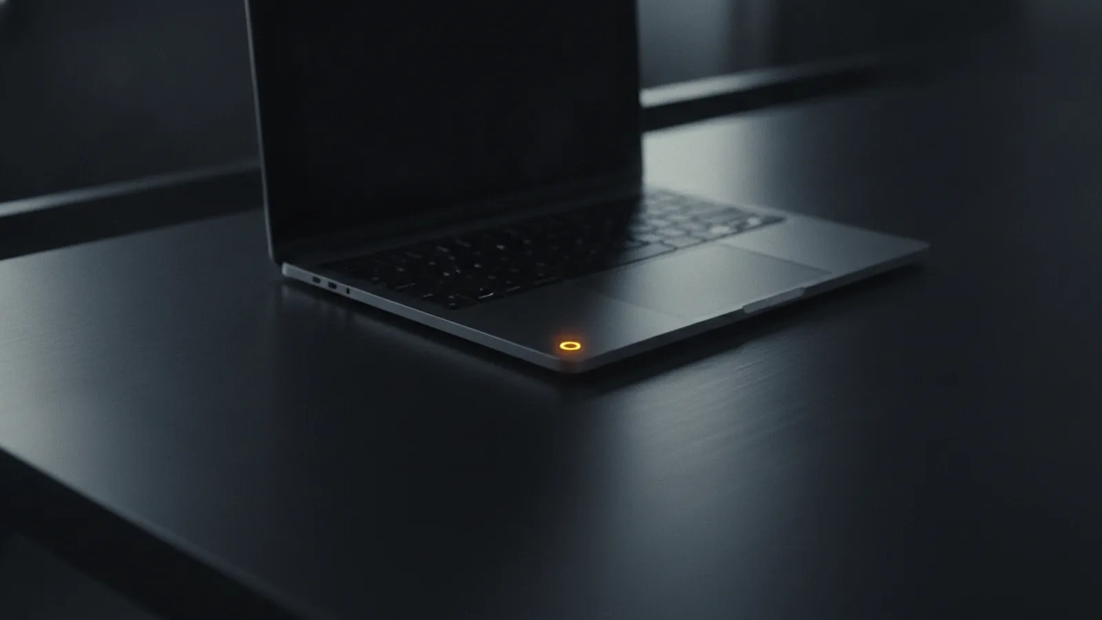 Un couvercle d’ordinateur portable refermé à moitié sur un bureau sombre et vide, sa seule diode d’alimentation ambre encore allumée tandis que l’écran s’éteint.